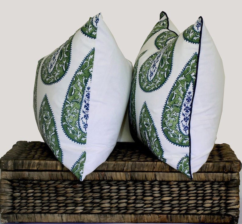 May include: Two white throw pillows with a green paisley pattern and a navy blue trim. The pillows are sitting on a brown wicker basket.