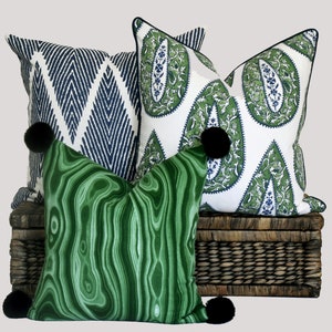 May include: Three decorative throw pillows with black pom-poms. The pillows are on a woven wicker basket. One pillow has a green and white paisley pattern, one has a blue and white geometric pattern, and one has a green marbled pattern.
