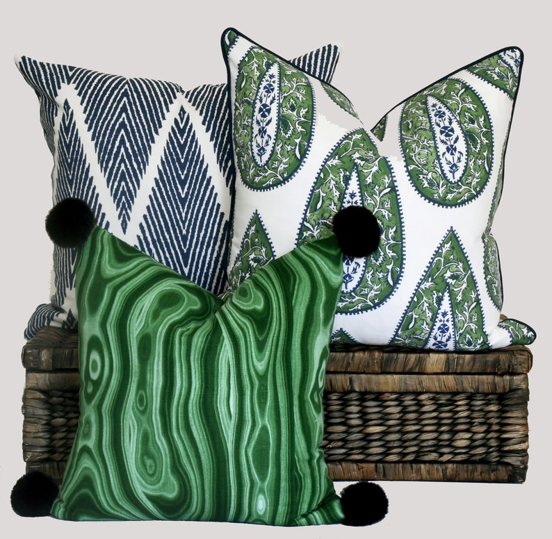 May include: Three decorative throw pillows with black pom-poms. The pillows are on a woven wicker basket. One pillow has a green and white paisley pattern, one has a blue and white geometric pattern, and one has a green marbled pattern.