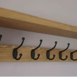 May include: A wall-mounted wooden coat rack featuring a natural light brown wood grain finish with a top shelf and ten evenly spaced black metal double hooks attached to the front board, supported by two side brackets on either end.