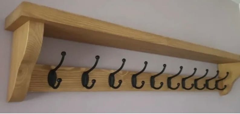 May include: A wall-mounted wooden coat rack featuring a natural light brown wood grain finish with a top shelf and ten evenly spaced black metal double hooks attached to the front board, supported by two side brackets on either end.