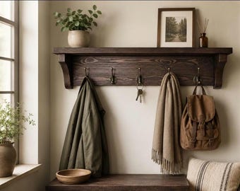 May include: A dark brown wooden wall-mounted shelf featuring five metal hooks. The shelf holds a potted plant and a framed picture. Hanging from the hooks are an olive green jacket, a set of keys, a beige fringed scarf, and a brown canvas backpack.