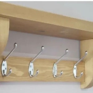 May include: A light-colored wooden wall shelf featuring a flat top surface and two curved side supports. The unit includes a horizontal back panel equipped with four evenly spaced, silver-toned metal coat hooks with double prongs.