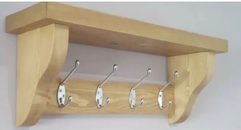 May include: A light-colored wooden wall shelf featuring a flat top surface and two curved side supports. The unit includes a horizontal back panel equipped with four evenly spaced, silver-toned metal coat hooks with double prongs.