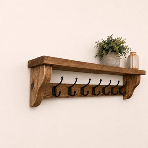 May include: A wall-mounted wooden shelf with a dark brown stained finish and curved side supports. The shelf features seven black metal hooks attached to the front panel. A small potted green plant and a beige ceramic vase sit on the top surface.