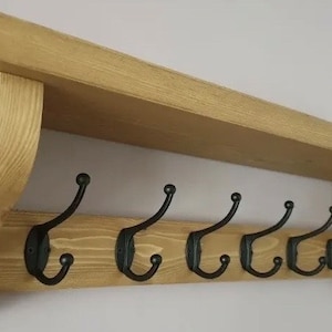 May include: A wall-mounted wooden shelf featuring a natural light brown wood grain finish and two curved side supports. The unit includes a horizontal backboard equipped with six evenly spaced black metal double hooks for hanging items.