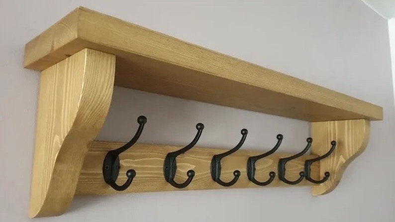 May include: A wall-mounted wooden shelf featuring a natural light brown wood grain finish and two curved side supports. The unit includes a horizontal backboard equipped with six evenly spaced black metal double hooks for hanging items.