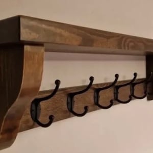 May include: A wall-mounted wooden shelf featuring a dark brown stained finish with a flat top surface and two side support brackets. Attached to the front board are five black metal hooks, each with a curved design for hanging various items.