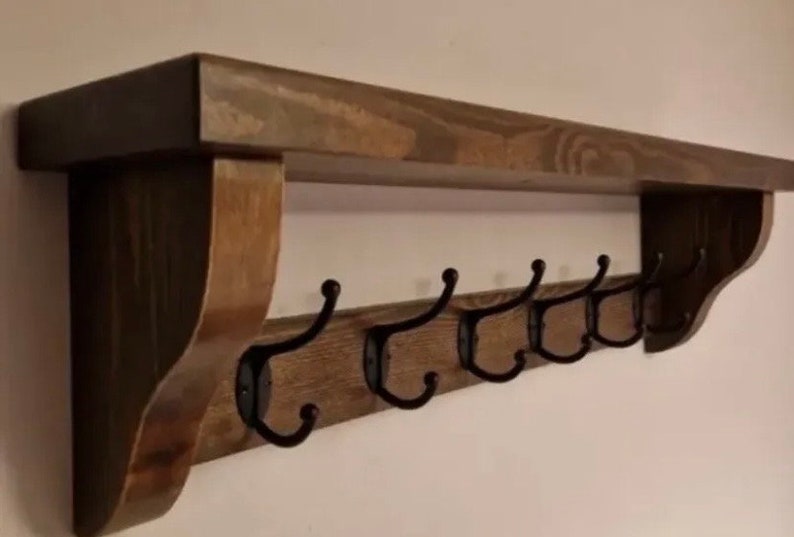 May include: A wall-mounted wooden shelf featuring a dark brown stained finish with a flat top surface and two side support brackets. Attached to the front board are five black metal hooks, each with a curved design for hanging various items.