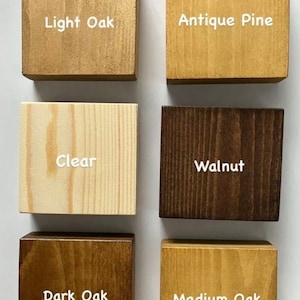 May include: Six square wooden blocks arranged in two columns, each displaying a different wood stain finish with white text labels: Light Oak, Antique Pine, Clear, Walnut, Dark Oak, and Medium Oak. Each block shows natural wood grain patterns.