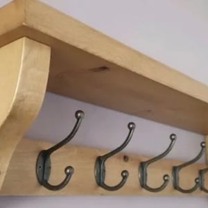 May include: A light-colored wooden wall shelf featuring a flat top surface and a lower horizontal board. Five dark metal double hooks are attached to the lower board, each with a curved upper arm and a smaller curved lower hook for hanging items.