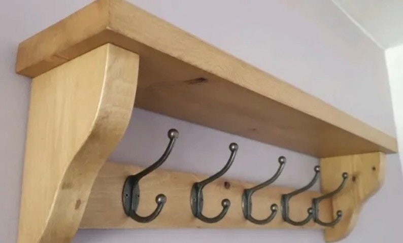 May include: A light-colored wooden wall shelf featuring a flat top surface and a lower horizontal board. Five dark metal double hooks are attached to the lower board, each with a curved upper arm and a smaller curved lower hook for hanging items.