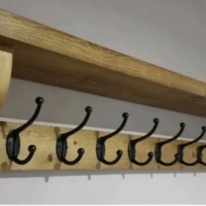 May include: A wall-mounted wooden coat rack featuring a natural light brown wood grain finish with a top shelf and two decorative side brackets. The rack includes nine black metal double hooks arranged in a horizontal row for hanging items.