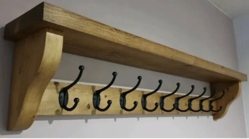 May include: A wall-mounted wooden coat rack featuring a natural light brown wood grain finish with a top shelf and two decorative side brackets. The rack includes nine black metal double hooks arranged in a horizontal row for hanging items.