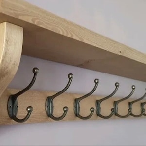 May include: A wall-mounted wooden shelf featuring a natural light brown wood grain finish and two curved side supports. Attached to the front board are nine dark metal coat hooks, each with a double-pronged design and rounded ball tips.