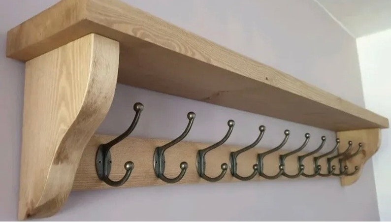 May include: A wall-mounted wooden shelf featuring a natural light brown wood grain finish and two curved side supports. Attached to the front board are nine dark metal coat hooks, each with a double-pronged design and rounded ball tips.