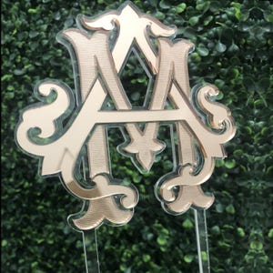May include: Gold mirrored acrylic monogram with the letters "A" and "M" intertwined. The letters are in a decorative script font and are mounted on a clear acrylic stand.