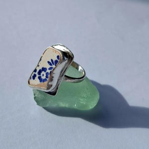 Könnte beinhalten: Ein Silberring mit einem rechteckigen Stück weißer Keramik als Herzstück, das mit einem blauen Blumenmuster verziert ist. Der Ring liegt auf einem durchscheinenden, hellgrünen, glatten und abgerundeten Stück Meerglas mit matter Textur.