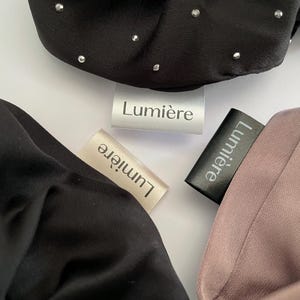 May include: Close-up of three satin scrunchies in black, beige, and mauve. The black scrunchie has small silver embellishments. Each scrunchie has a tag that reads "Lumière".