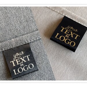 K&ouml;nnte beinhalten: Zwei schwarze Stoff-Etiketten mit goldener Aufschrift "Ihr Text oder Logo" auf einem grauen und beigefarbenen Stoffhintergrund.