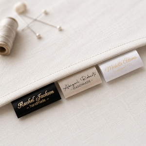 Puede incluir: Tres etiquetas rectangulares de tela para ropa unidas a un tejido color crema. Una etiqueta negra con texto dorado Rachel Jackson - handmade, una etiqueta beige con texto negro Abigail Roberts - HANDMADE y una etiqueta blanca con texto dorado.