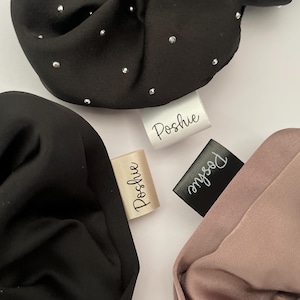 May include: Close-up of three fabric hair scrunchies in black, tan, and mauve. The black scrunchie has small silver embellishments. Each scrunchie has a tag that reads "Poshie" in a handwritten font.