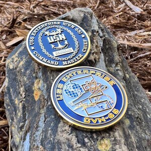 May include: Two gold-colored challenge coins with blue and white designs. The top coin features a navy anchor and the text "USS ENTERPRISE (CVN 65) ★ USNS COMMAND MASTER CHIEF". The bottom coin features a stylized image of a spaceship and the text "USS ENTERPRISE CVN-65".