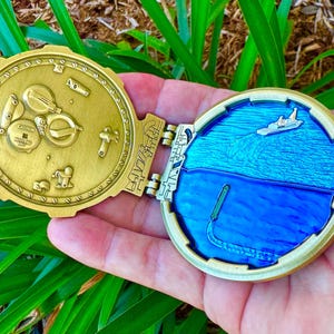 May include: A detailed, open, gold-colored, hinged medallion. One side features a blue ocean scene with a boat, the other side has detailed mechanical elements. The medallion is held in a hand against a background of green leaves.