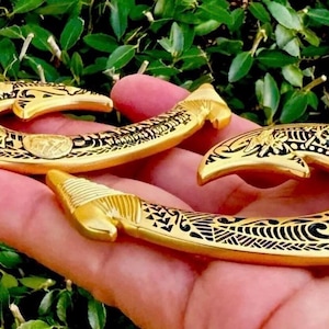 May include: Three gold-colored, hook-shaped pendants with intricate black etched designs. The pendants are held in a hand against a backdrop of green foliage. The hooks have a curved shape with detailed patterns.
