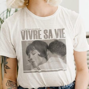 Puede incluir: Camiseta blanca con una foto en blanco y negro de una pareja y el texto "VIVRE SA VIE" impreso en la parte delantera.
