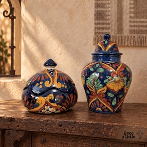 May include: Two hand-painted ceramic vessels with lids, featuring a glossy finish in deep blue, yellow, and terracotta red. The shorter, rounded bowl and the taller, urn-shaped jar both display intricate floral and geometric patterns throughout.