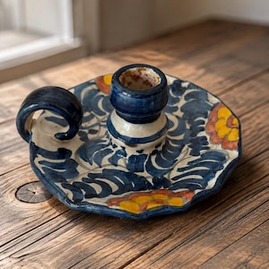 May include: A hand-painted ceramic candle holder with a scalloped edge, featuring a dark blue leaf pattern on a cream background with yellow and orange floral accents. It includes a central cylindrical candle socket and a curved side handle.