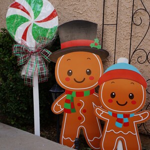 May include: Christmas yard decorations featuring a large lollipop and gingerbread man cutouts. The lollipop has red, white, and green swirls. The gingerbread men wear hats and scarves. The decorations are on white stakes.