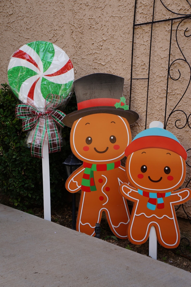 May include: Christmas yard decorations featuring a large lollipop and gingerbread man cutouts. The lollipop has red, white, and green swirls. The gingerbread men wear hats and scarves. The decorations are on white stakes.