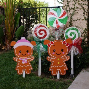 May include: Outdoor Christmas decorations featuring gingerbread people and candy-themed cutouts. The gingerbread figures have pink and white accents. Large, colorful lollipops with red, green, and white swirls are also displayed, adding a festive touch to the yard.