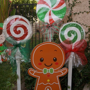 May include: Outdoor Christmas decorations featuring a gingerbread man and large, colorful lollipops. The lollipops have red and green swirls, wrapped in cellophane, and adorned with bows. The gingerbread man is orange with a green bow tie.