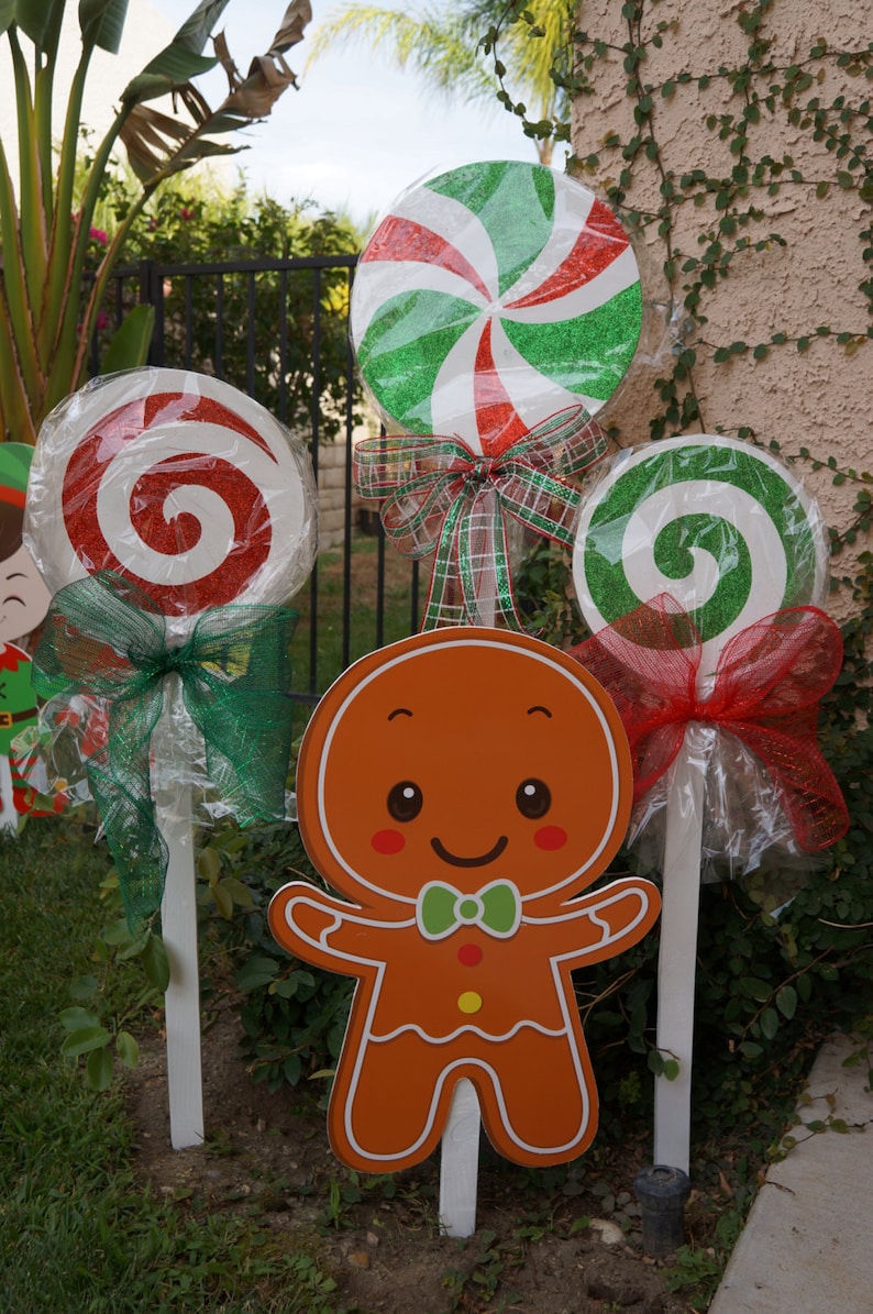 May include: Outdoor Christmas decorations featuring a gingerbread man and large, colorful lollipops. The lollipops have red and green swirls, wrapped in cellophane, and adorned with bows. The gingerbread man is orange with a green bow tie.