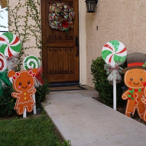 May include: Gingerbread man and candy cane yard decorations. The gingerbread men are wearing hats and scarves. The candy canes are red, white, and green. The decorations are on white stakes and are placed in the grass.
