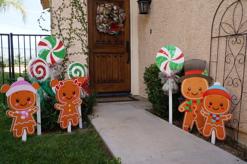 May include: Gingerbread man and candy cane yard decorations. The gingerbread men are wearing hats and scarves. The candy canes are red, white, and green. The decorations are on white stakes and are placed in the grass.