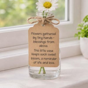 Op de afbeelding: Een kleine glazen vaas met een enkele madelief met een gele kern en witte bloemblaadjes. De vaas heeft een touwen strik en een label met de tekst: "Flowers gathered by tiny hands - blessings from above. This little vase keeps each sweet bloom, a reminder of life and love."
