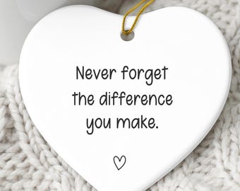 May include: A white heart-shaped ornament with the text "Never forget the difference you make" and a small heart symbol. A gold string is attached for hanging. A mug of hot chocolate with marshmallows and a cinnamon stick is in the background.