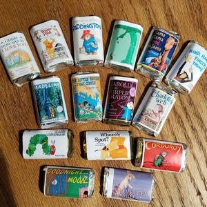 May include: A collection of wrapped chocolate bars featuring book covers. Titles include "Guess How Much I Love You," "Winnie the Pooh," "Paddington," and "The Very Hungry Caterpillar." The bars are arranged on a wooden surface.
