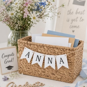 May include: A woven basket with a banner that spells "ANNA" is filled with cards. A small sign reads "Congrats Grad!" and a wooden cutout says "Graduate." A bouquet of flowers and blue ribbon complete the graduation theme.