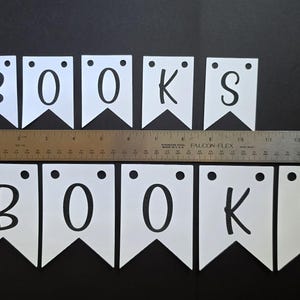Może przedstawiać: Dziesięć białych proporczyk&oacute;w z czarnymi literami tworzącymi dwukrotnie słowo BOOKS, każdy z dwoma okrągłymi otworami u g&oacute;ry. Metalowa linijka umieszczona poziomo pośrodku wskazuje całkowitą szerokość wynoszącą około 45,7 cm.