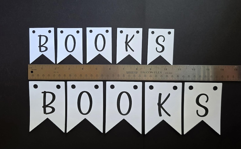 Może przedstawiać: Dziesięć białych proporczyk&oacute;w z czarnymi literami tworzącymi dwukrotnie słowo BOOKS, każdy z dwoma okrągłymi otworami u g&oacute;ry. Metalowa linijka umieszczona poziomo pośrodku wskazuje całkowitą szerokość wynoszącą około 45,7 cm.
