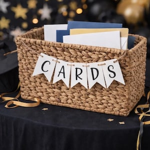 May include: A woven wicker basket filled with cards, adorned with a banner that reads "CARDS". The basket sits on a black surface, with gold ribbon and star-shaped confetti scattered around. The background features twinkling lights.