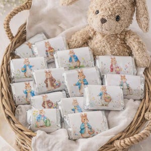 May include: A woven basket filled with individually wrapped chocolate bars featuring illustrations of Peter Rabbit. A plush bunny toy sits in the basket. The bars have colorful images of Peter Rabbit in various poses.