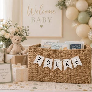 May include: A rectangular woven seagrass basket featuring a white pennant banner with the black text BOOKS. The basket contains several children's books. The scene includes a tan teddy bear, wrapped gifts, and a sign that reads THANK YOU.