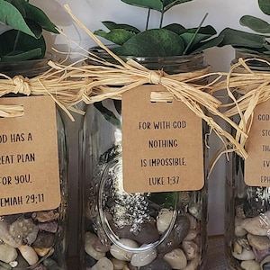 May include: Three glass jars with greenery, stones, and inspirational quotes on brown paper tags. The quotes are: "God has a great plan for you. Jeremiah 29:11", "For with God nothing is impossible. Luke 1:37", and "God has more in store for you than you can even imagine. Ephesians 3:20".