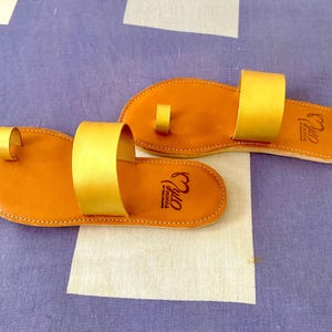 May include: A pair of brown leather sandals with gold straps. The sandals have a toe loop and the brand name "Myo" is printed on the straps.